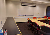 A ring of tables and chairs with a countertop to the left and a whiteboard and projector screen along back wall