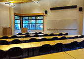 5 rows of chairs and tables lead forward to a podium, whiteboard, screen, and windows.